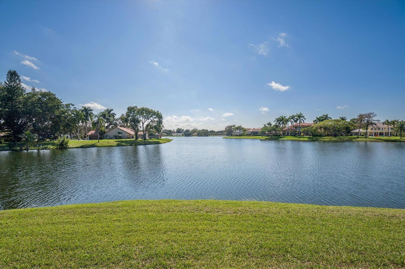A view of a lake surrounded by grassy lawns and residential houses under a sky — 7600 Hyannis Lane, Parkland FL 33067, PARKLAND, FL