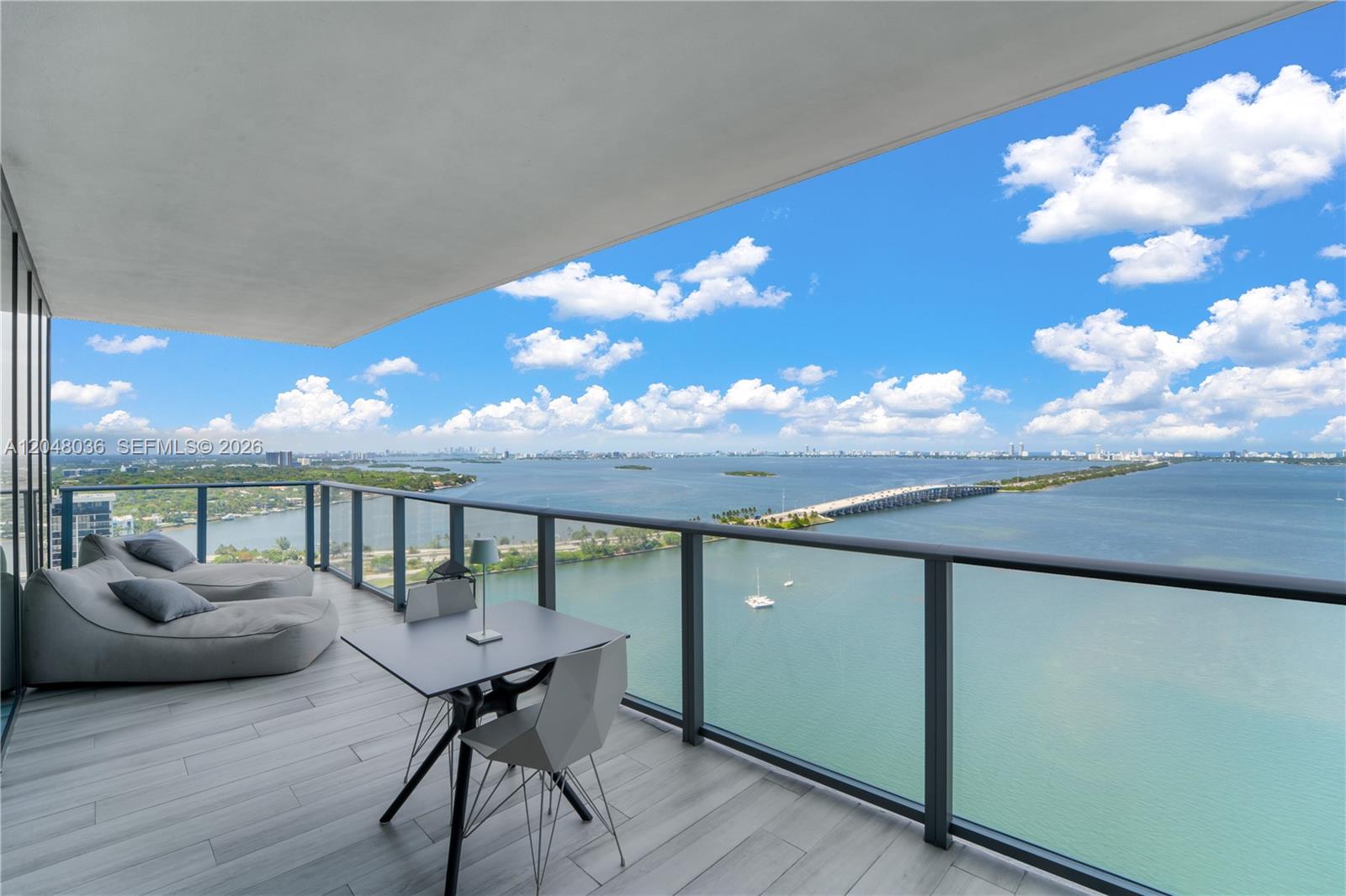 A high-rise balcony features gray lounge chairs, a small table, and panoramic water views — 3131 NE 7th Ave # 2706, Miami FL 33137, MIAMI, FL