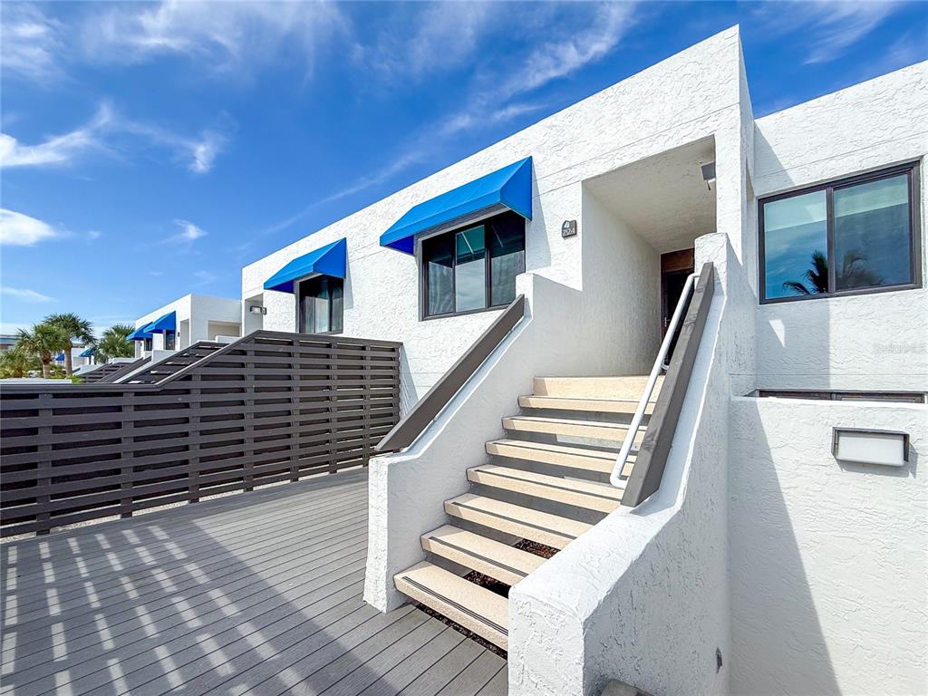 A white stucco building exterior features a wooden deck, a staircase, and blue window awnings — 724 BAYPORT WAY UNIT 724, LONGBOAT KEY, FL