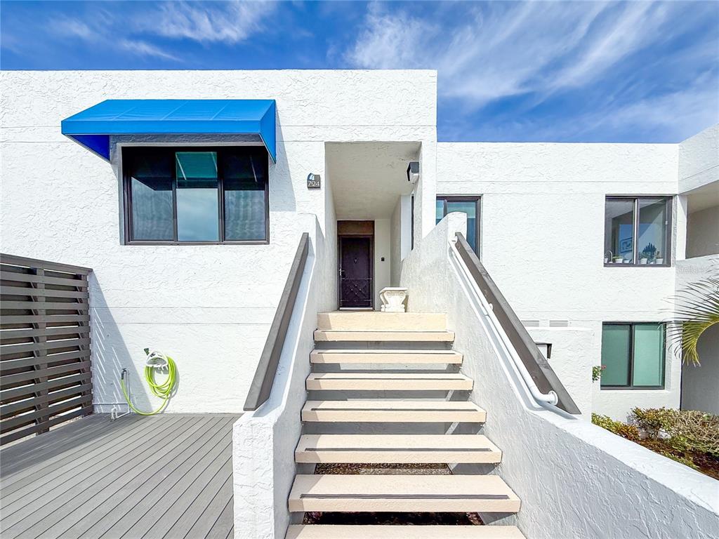 A white stucco building exterior features a blue awning, a wooden deck, and concrete stairs — 724 BAYPORT WAY UNIT 724, LONGBOAT KEY, FL