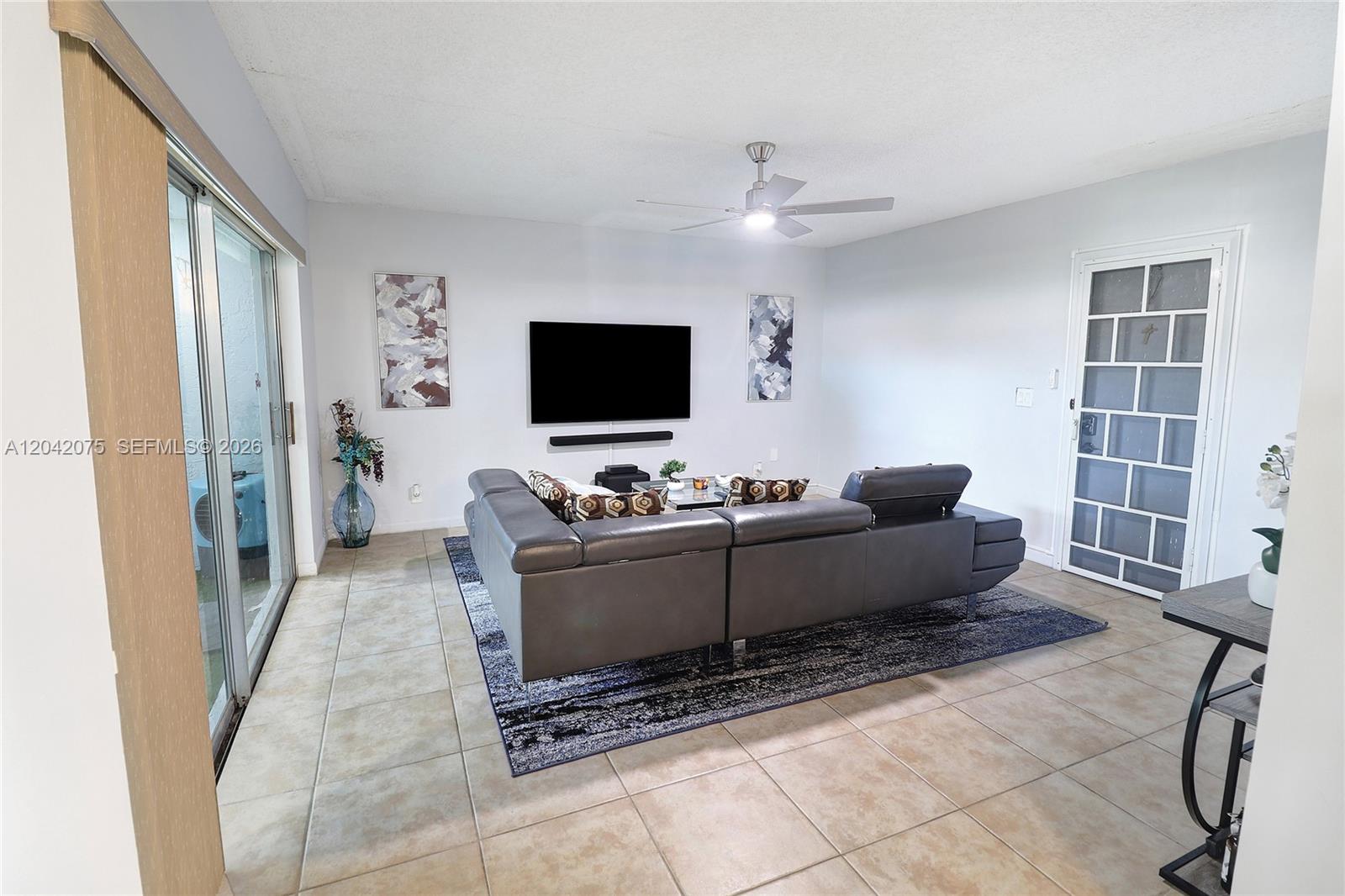 A living room features a gray sectional sofa, a wall-mounted television, and tile flooring — 14771 Wildflower Ln, Delray Beach FL 33446, DELRAY BEACH, FL