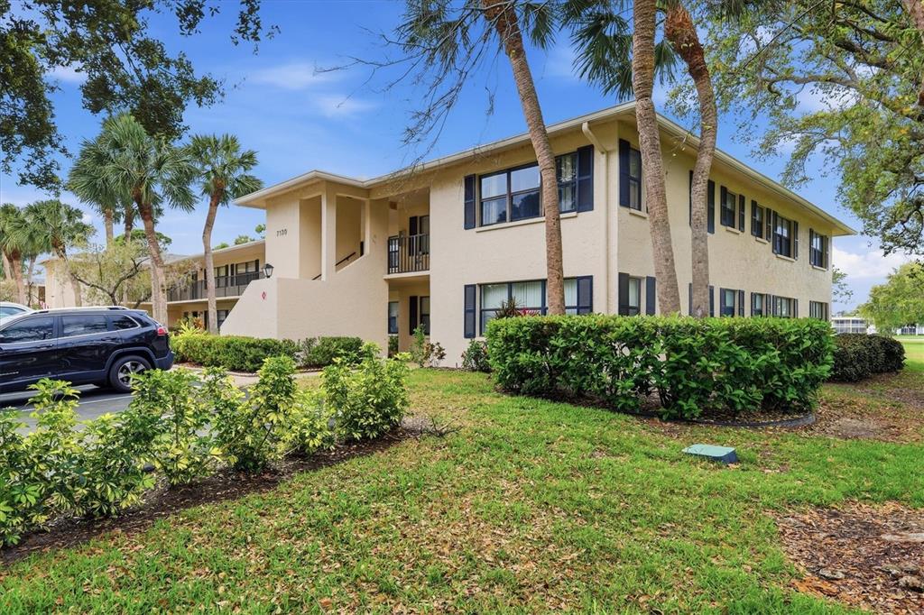 A two-story apartment building with beige stucco walls and dark shutters sits near palm trees — 7130 FAIRWAY BEND LANE UNIT 291, SARASOTA, FL
