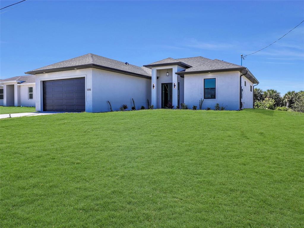 A single-story house with a white stucco exterior, a dark garage door, and a grassy lawn — 1310 NW 11TH TERRACE, CAPE CORAL, FL