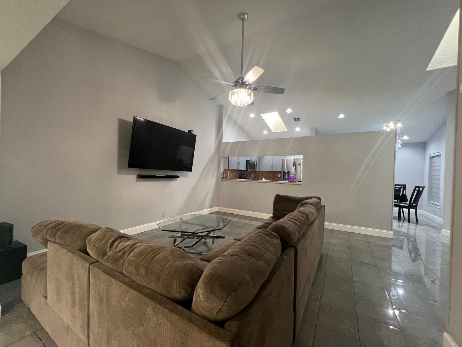 A living room with a brown sectional, glass coffee table, wall-mounted television, and tile flooring — 4241 Green Forest Way, Boynton Beach FL 33436, BOYNTON BEACH, FL