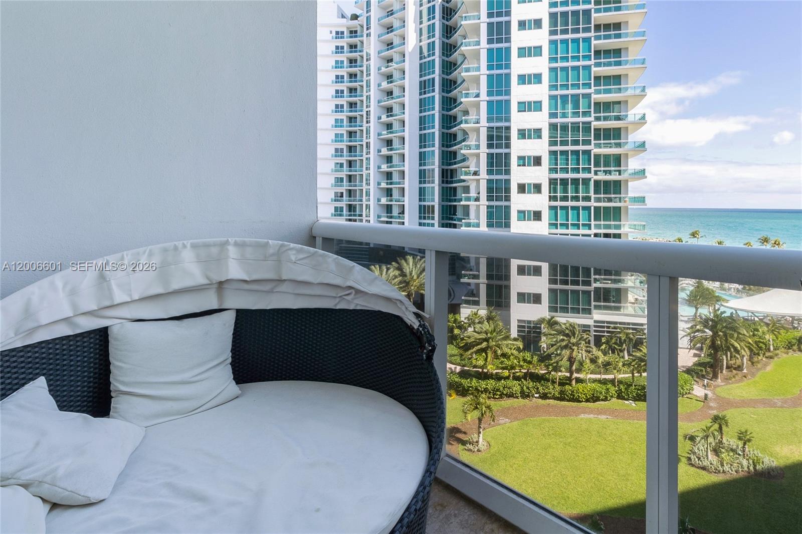 A balcony features a round outdoor daybed overlooking a grassy lawn, palm trees, and ocean — BAL HARBOUR, FL