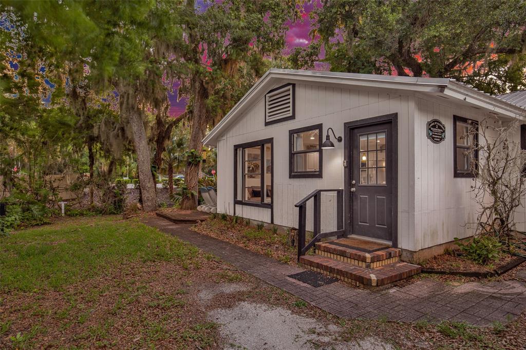 A white cottage with dark trim and a brick-stepped entrance sits in a wooded yard — 801 CANAL STREET, NEW SMYRNA BEACH, FL