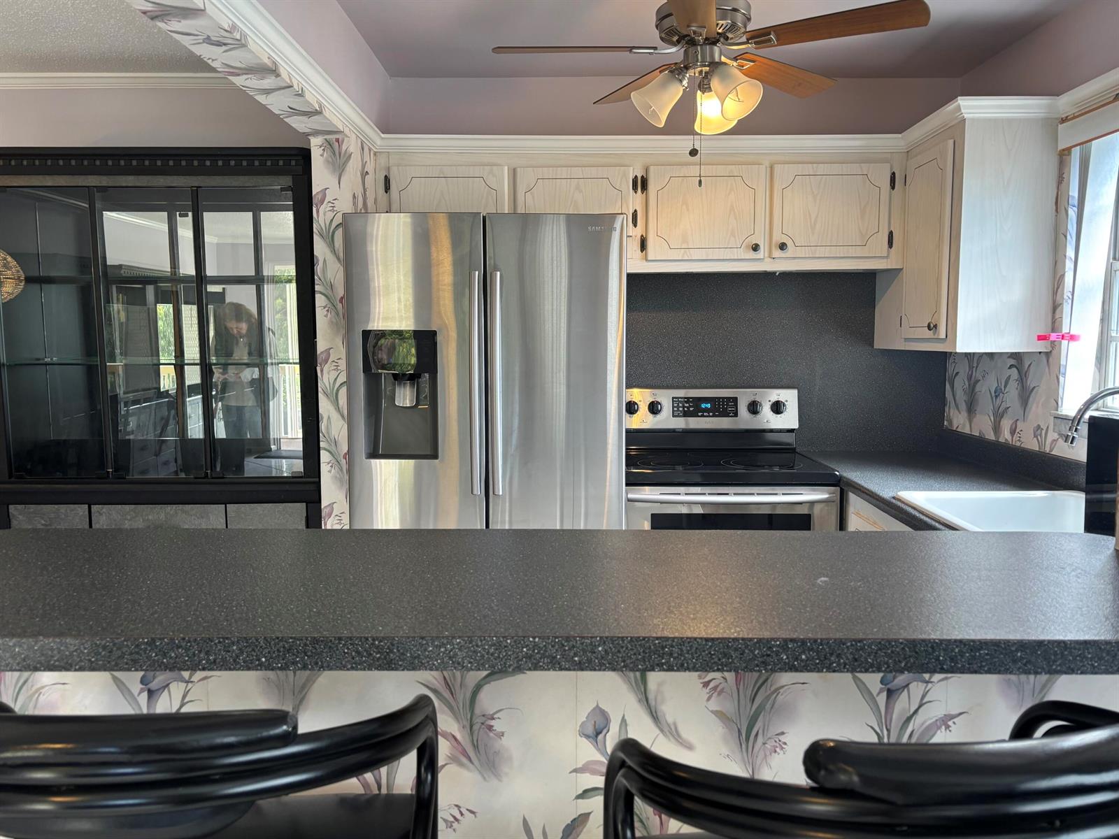 A kitchen features a stainless steel refrigerator, dark countertops, floral wallpaper, and a ceiling fan — 81 Windsor D, West Palm Beach FL 33417, WEST PALM BEACH, FL