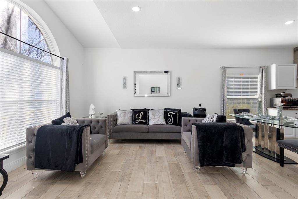 A living room features gray tufted sofas, light wood-look flooring, and large windows with blinds — 1739 CANOE CREEK FALLS DRIVE, ORLANDO, FL