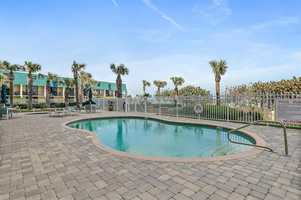 An outdoor swimming pool with a paved deck is surrounded by a fence and palm trees — 711 S ATLANTIC AVENUE Unit 202, NEW SMYRNA BEACH, FL