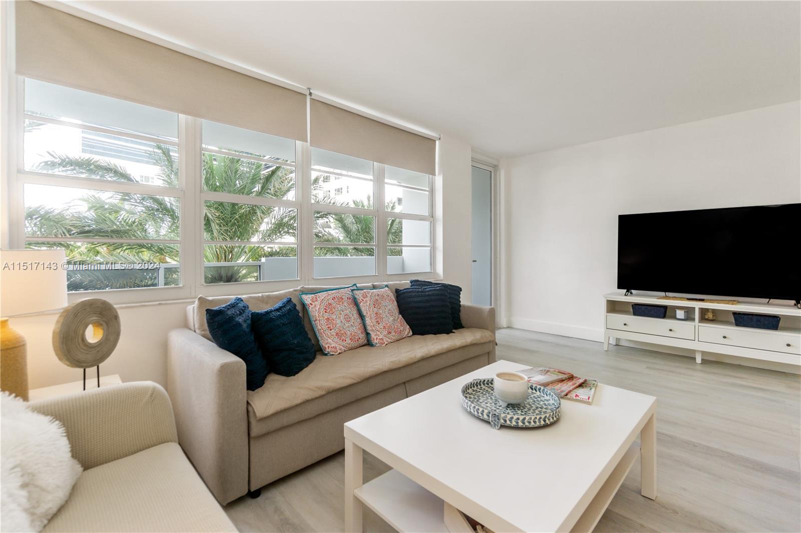 A living room features a beige sofa, white coffee table, and large windows with palm trees — 100 Lincoln Rd # 312, Miami Beach FL 33139, MIAMI BEACH, FL