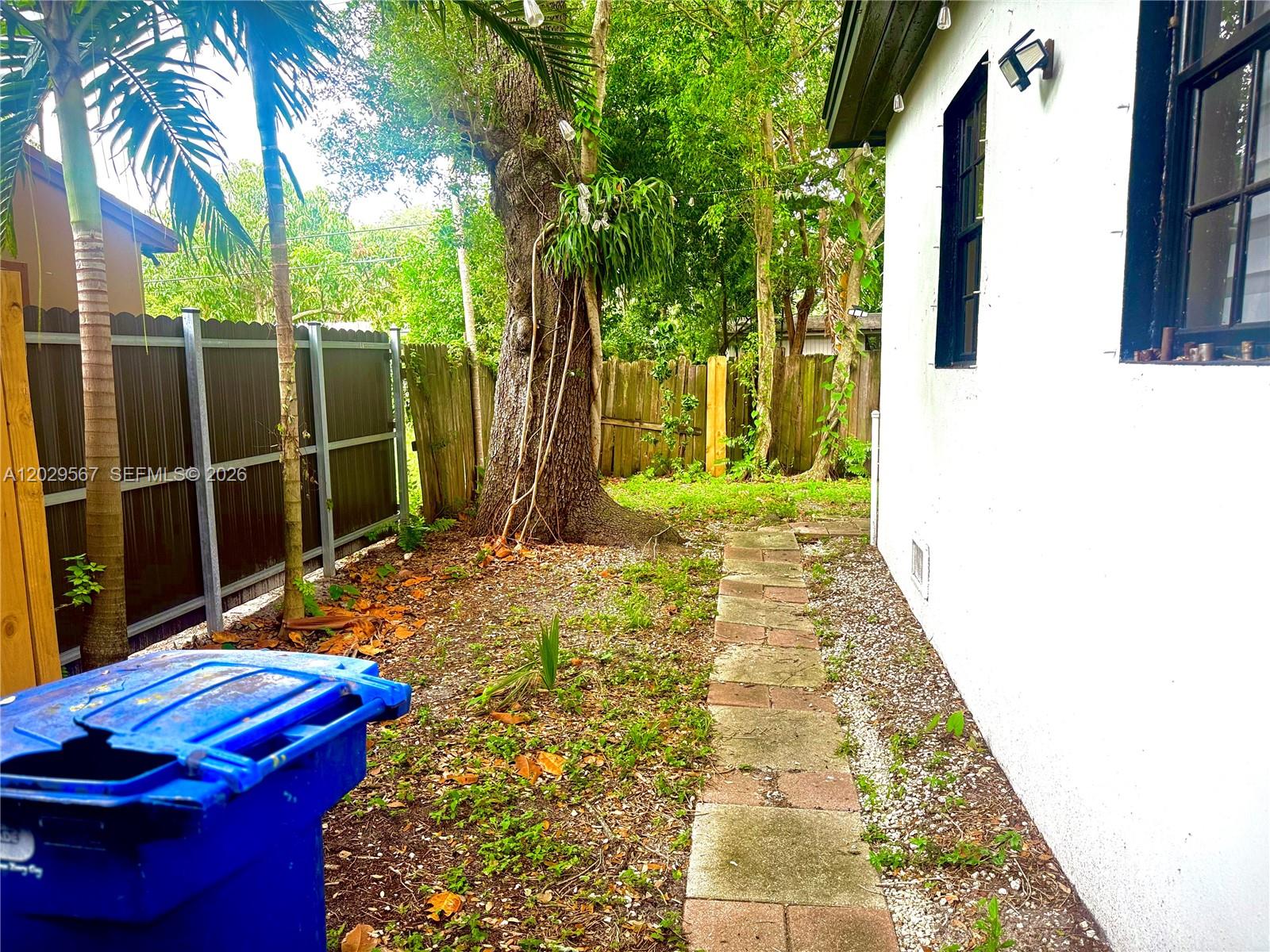 A narrow stone walkway runs alongside a white house wall toward a large tree — 620 NE 160th St, Miami FL 33162, MIAMI, FL