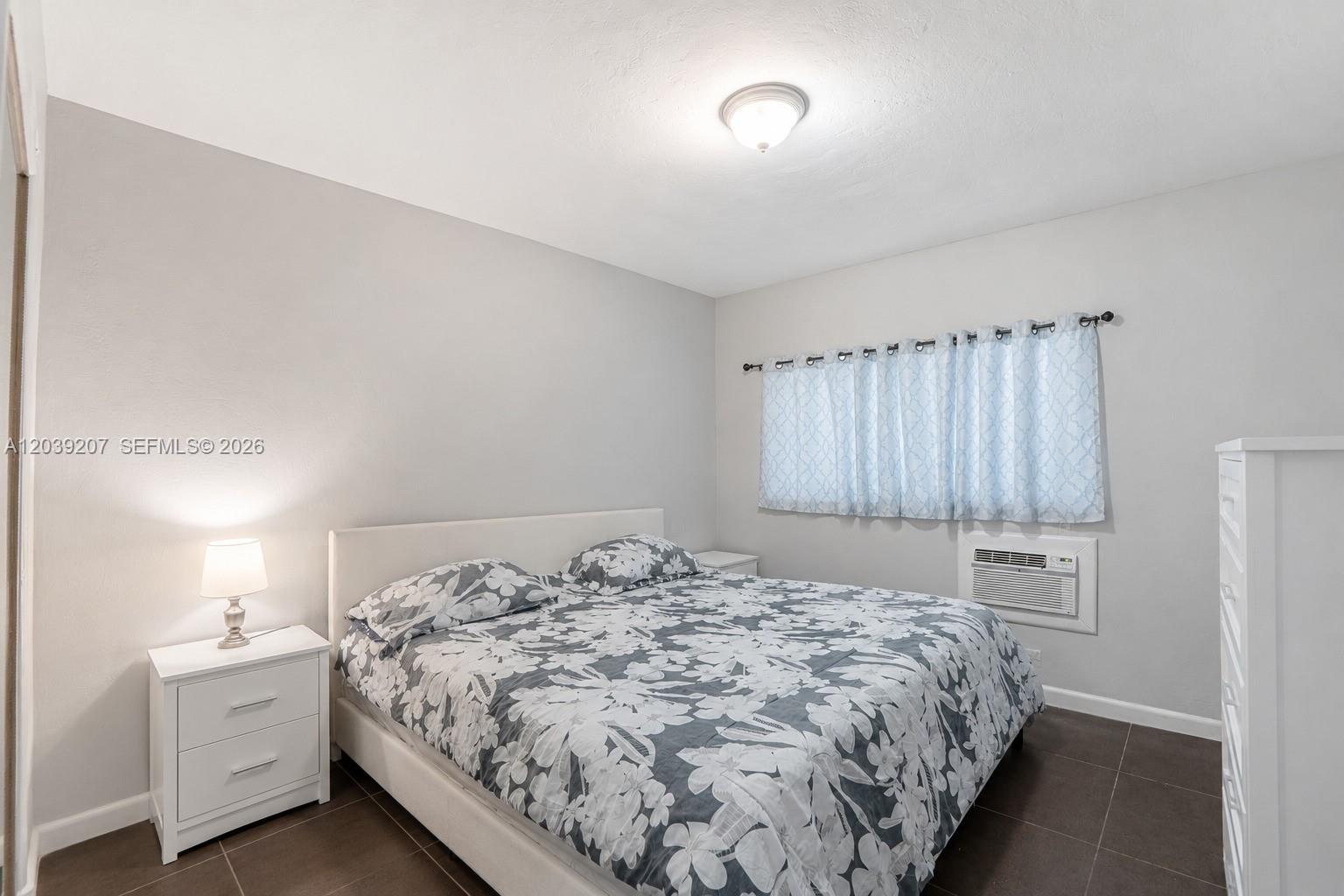 A bedroom features a floral-patterned bed, white nightstands, tile flooring, and a window air conditioner — 521 NE 82nd Ter # 3, Miami FL 33138, MIAMI, FL