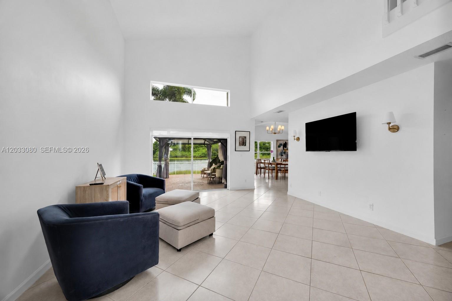 A bright living room features high ceilings, tile floors, blue armchairs, and a wall-mounted television — 18018 SW 30th Ct, Miramar FL 33029, MIRAMAR, FL