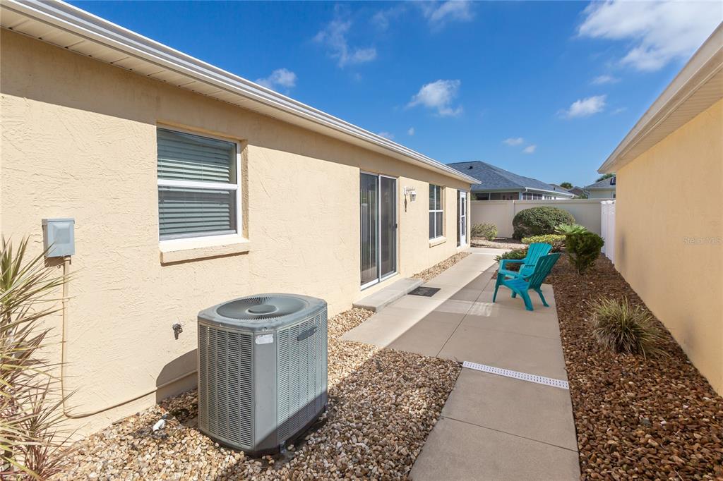 A beige house exterior features a concrete patio, an air conditioning unit, and a blue chair — 1497 ALEXANDRIA PLACE, THE VILLAGES, FL