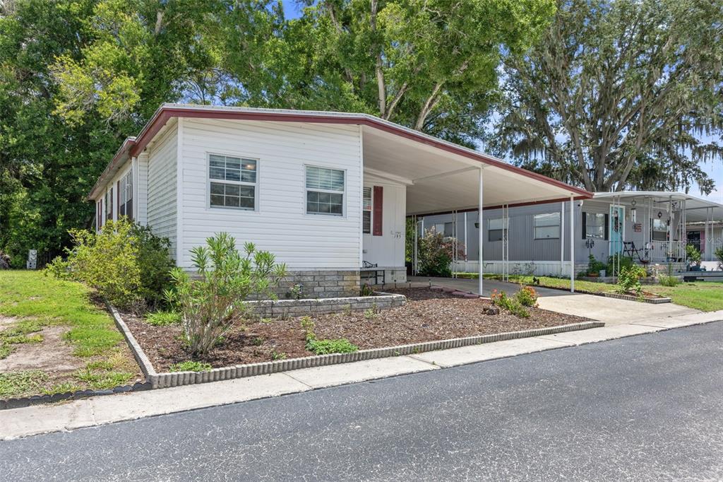 A white mobile home with a carport and a landscaped front yard sits along a road — 15601 LAKESHORE VILLA LOOP, TAMPA, FL