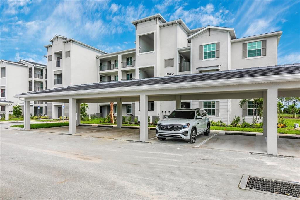A modern multi-story apartment building features a covered parking area with a parked SUV — 12320 WELLEN GOLF STREET UNIT 204, VENICE, FL