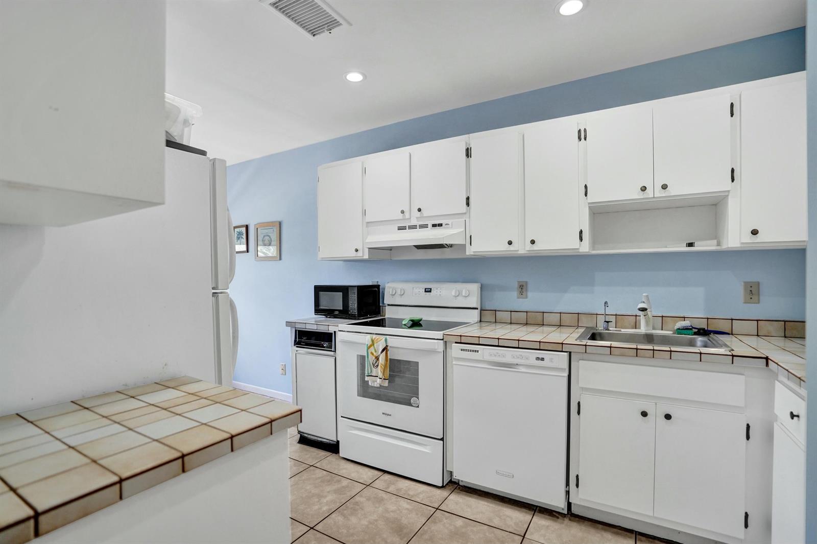 A kitchen features white cabinets, a white stove, a dishwasher, and tan tiled countertops — 19 Stratford Drive E Unit F, Boynton Beach FL 33436, BOYNTON BEACH, FL