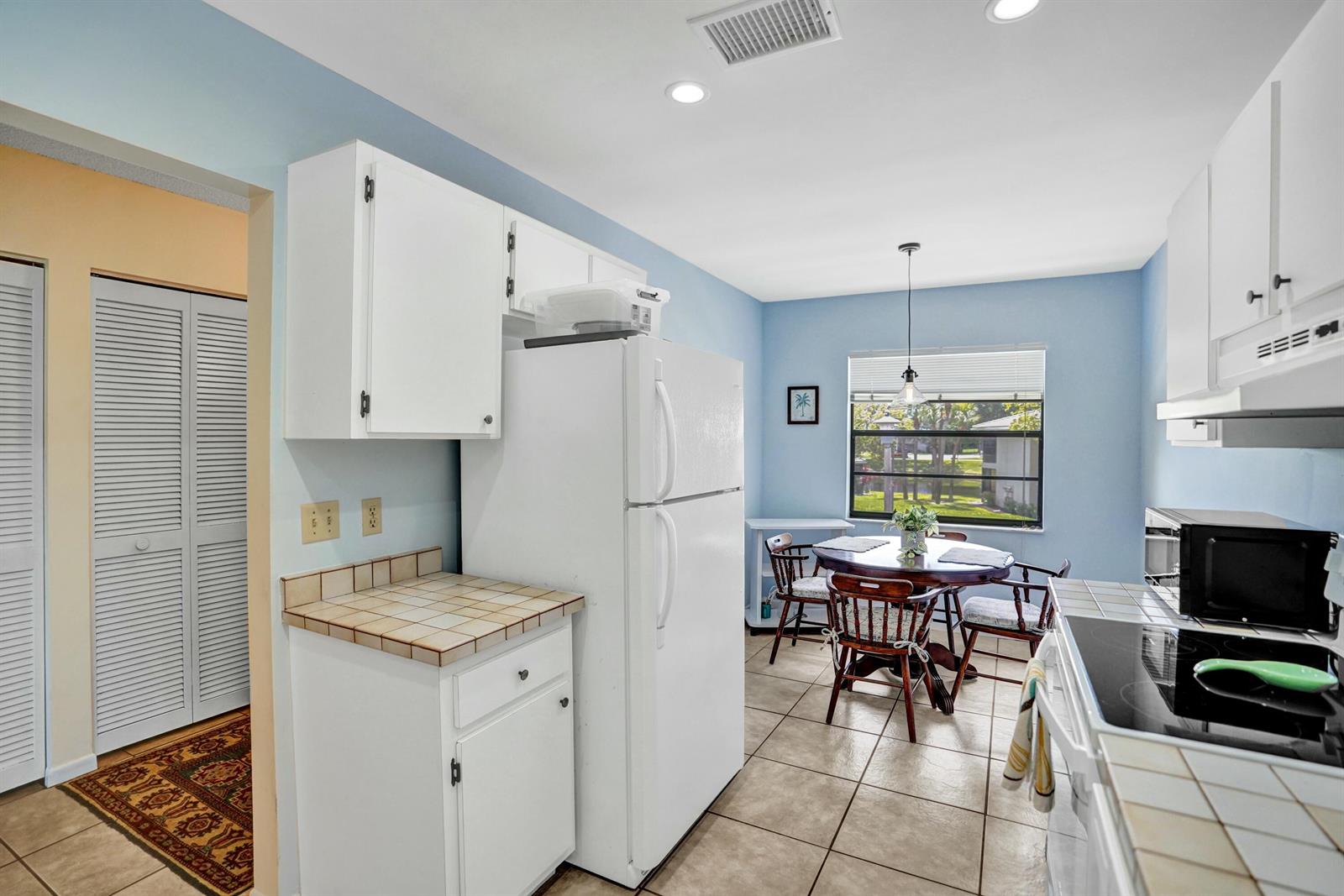 A kitchen with white cabinets, tile countertops, a white refrigerator, and a small dining area — 19 Stratford Drive E Unit F, Boynton Beach FL 33436, BOYNTON BEACH, FL