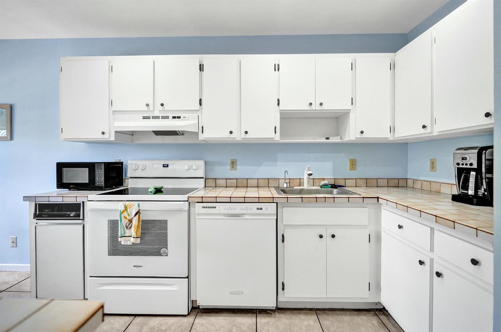 A kitchen with white cabinets, white appliances, and light-colored tiled countertops against blue walls — 19 Stratford Drive E Unit F, Boynton Beach FL 33436, BOYNTON BEACH, FL
