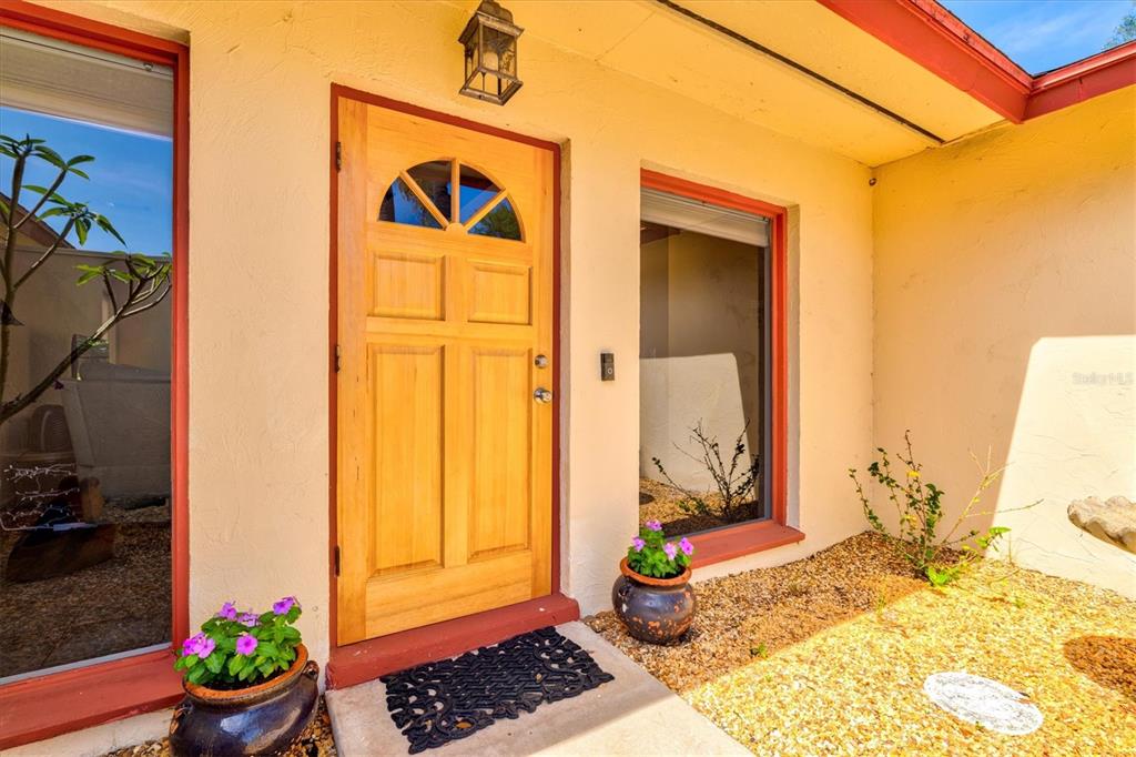 A light-colored wooden front door is flanked by two large windows and two potted plants — 6533 BOWLINE DRIVE, SARASOTA, FL