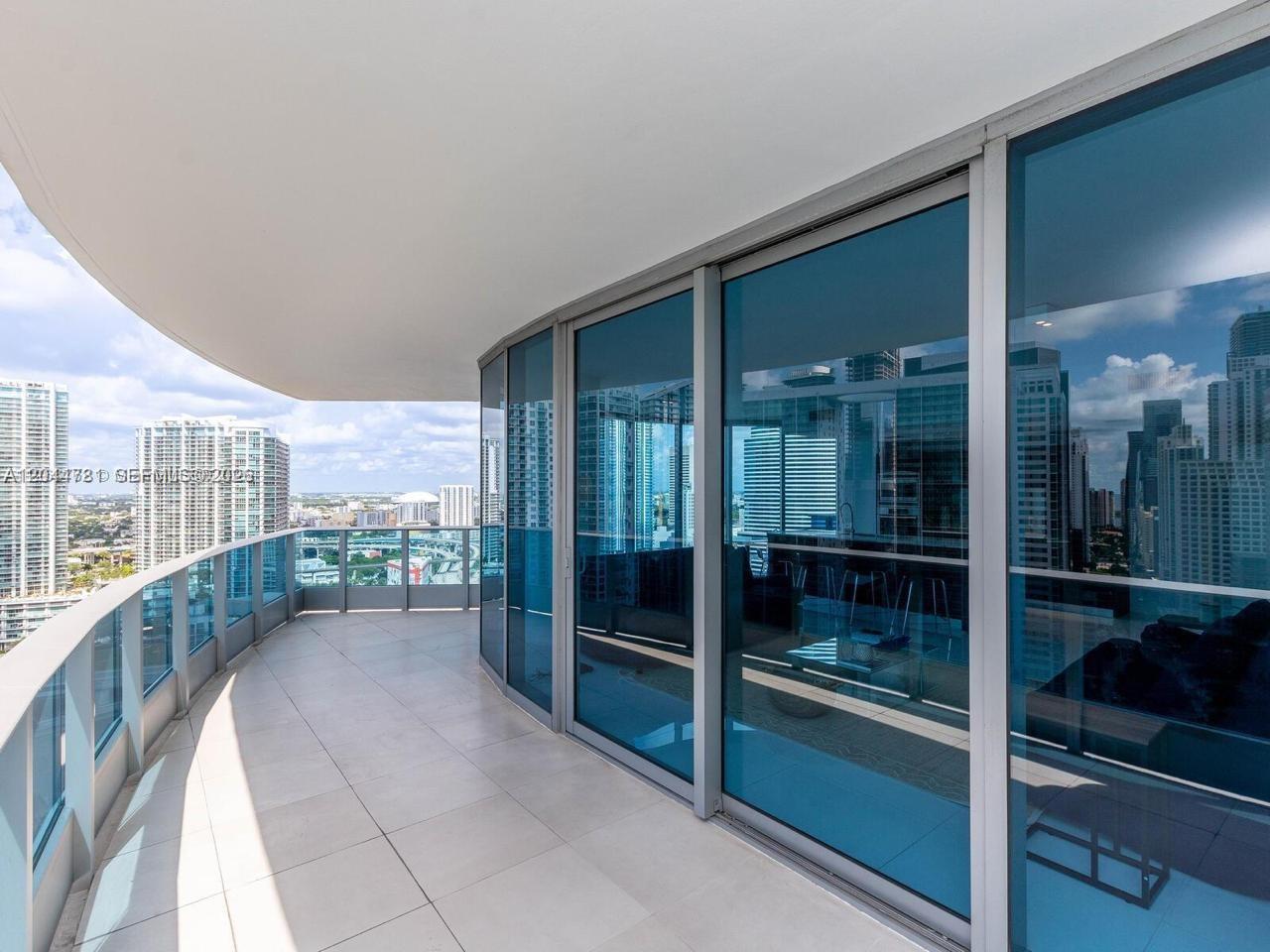 A curved balcony with light-colored tile flooring overlooks a city skyline through glass railings — 200 Biscayne Boulevard Way # 3102, Miami FL 33131, MIAMI, FL
