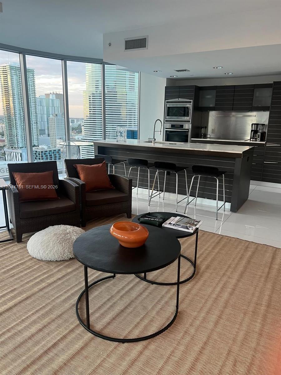 A modern apartment living area features a kitchen island, dark cabinetry, and floor-to-ceiling windows — 200 Biscayne Boulevard Way # 3102, Miami FL 33131, MIAMI, FL
