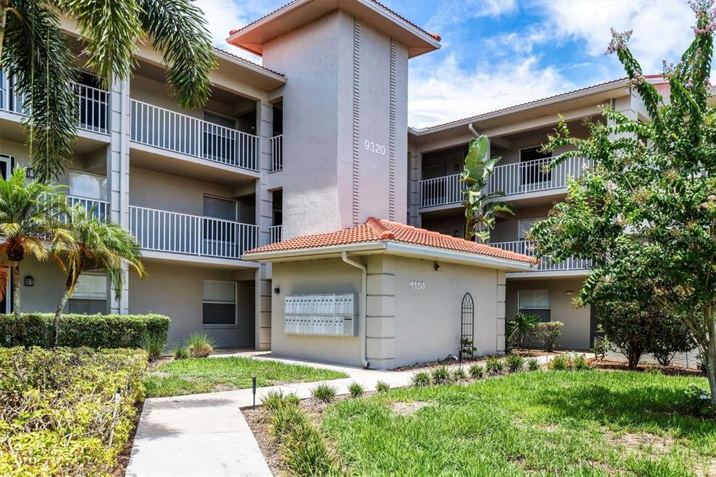 A three-story apartment building exterior features a central mail kiosk and a concrete walkway — 9320 CLUBSIDE CIRCLE UNIT 2108, SARASOTA, FL