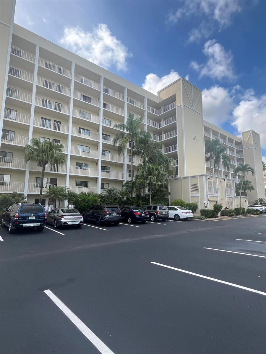 A multi-story residential building with balconies stands behind a paved parking lot — 7310 Ashford Place # 401, Delray Beach FL 33446, DELRAY BEACH, FL