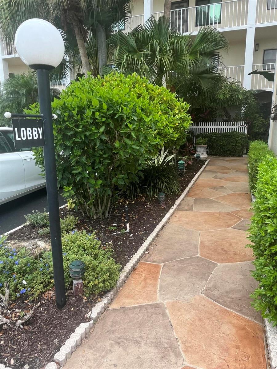 A stone walkway leads toward a building entrance marked by a lobby sign and landscaping — 7310 Ashford Place # 401, Delray Beach FL 33446, DELRAY BEACH, FL