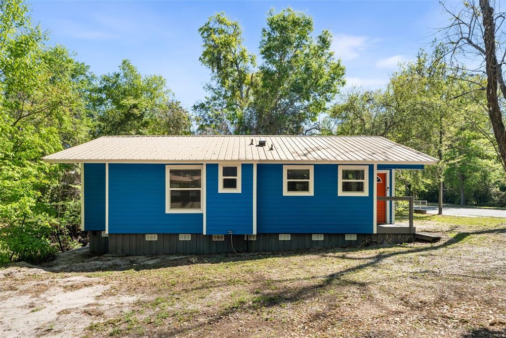 A blue, single-story house with a metal roof sits on a raised foundation among trees — 2256 W UNIVERSITY AVENUE, GAINESVILLE, FL