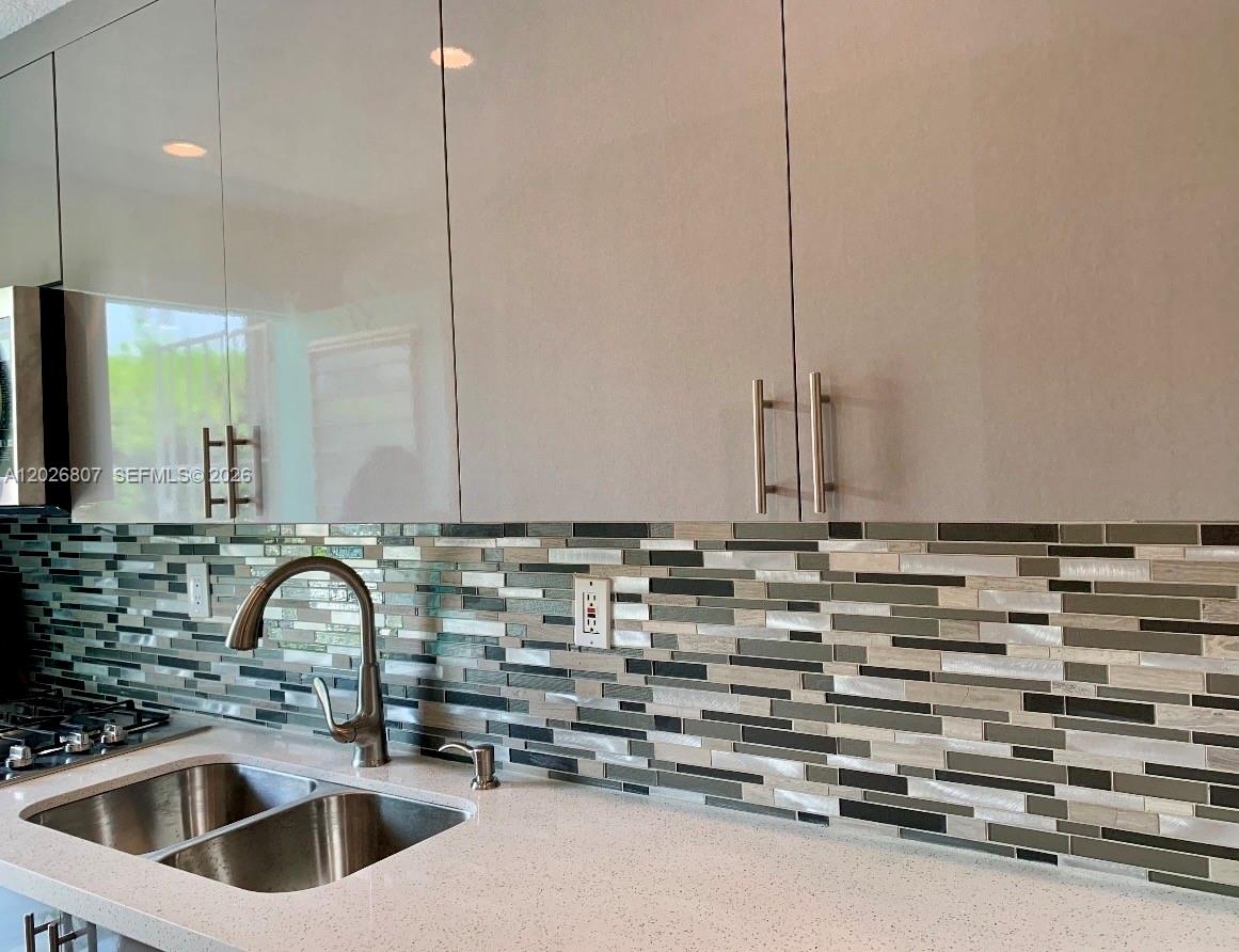 A kitchen features glossy light gray cabinets, a multi-colored glass tile backsplash, and a stainless steel sink — 6498 Collins Ave # 41, Miami Beach FL 33141, MIAMI BEACH, FL