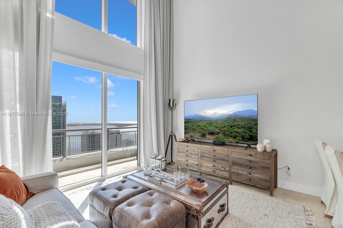 A living room features a large window, a television on a wooden dresser, and ottomans — 60 SW 13th St # 3402, Miami FL 33130, MIAMI, FL