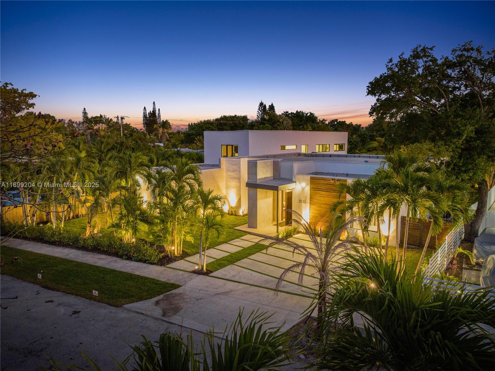 An exterior view of a modern white house with a wood-paneled entrance and palm trees — 2110 NE 197th Ter, Miami FL 33179, MIAMI, FL