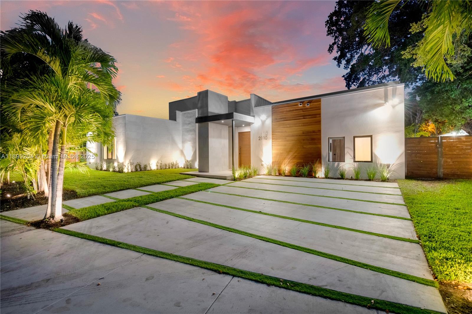 A modern single-story house with a concrete driveway and wood-paneled facade at sunset — 2110 NE 197th Ter, Miami FL 33179, MIAMI, FL