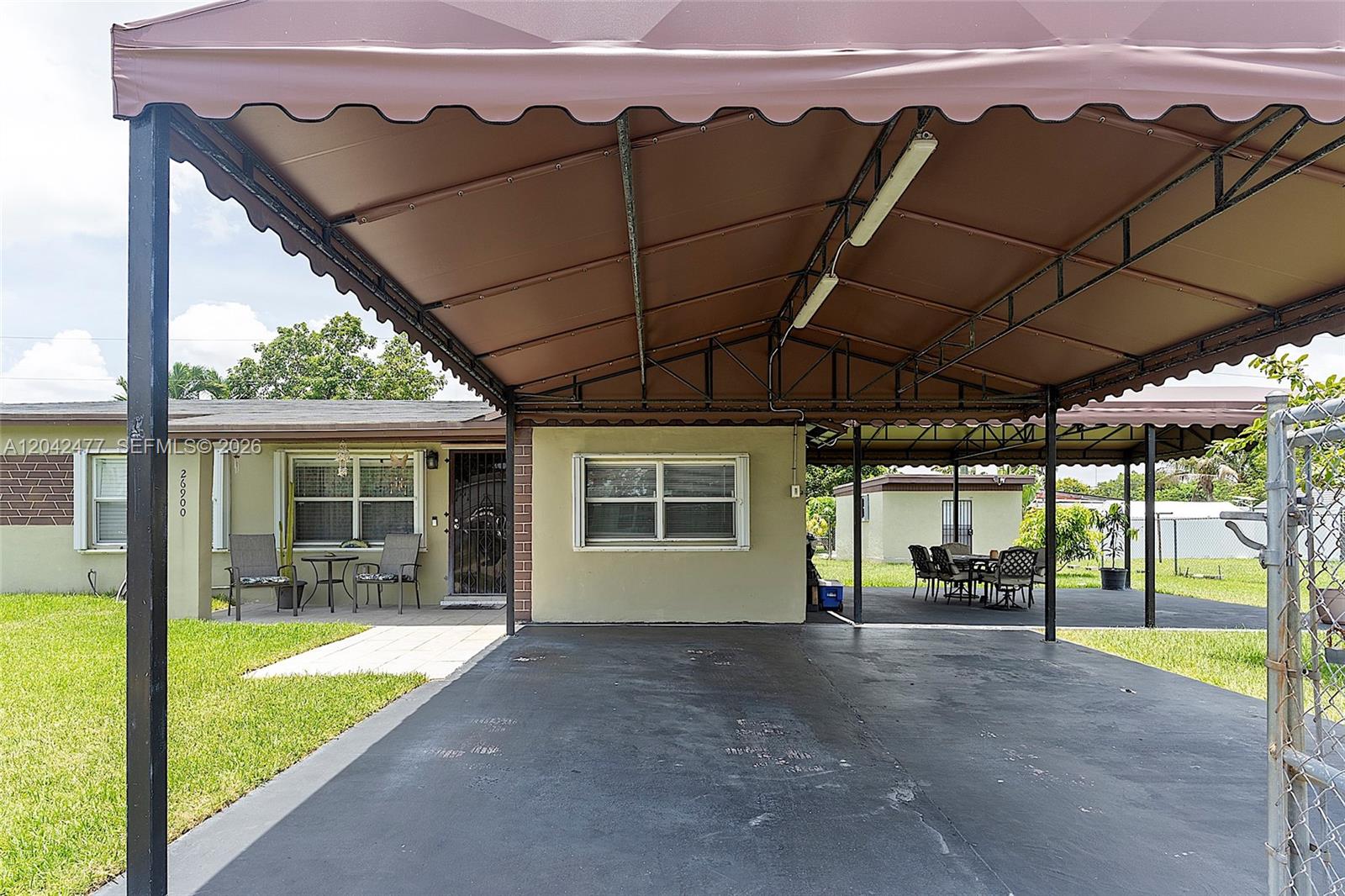 A house exterior features a large brown metal carport covering a paved driveway and patio — HOMESTEAD, FL