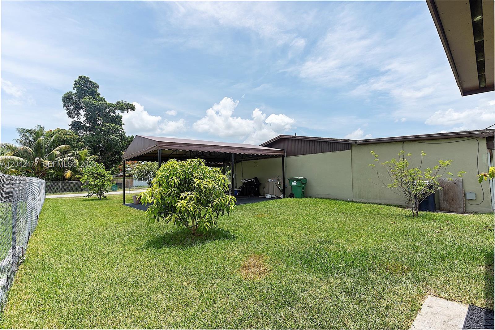 A grassy backyard features a metal carport, a small tree, and a chain-link fence — HOMESTEAD, FL