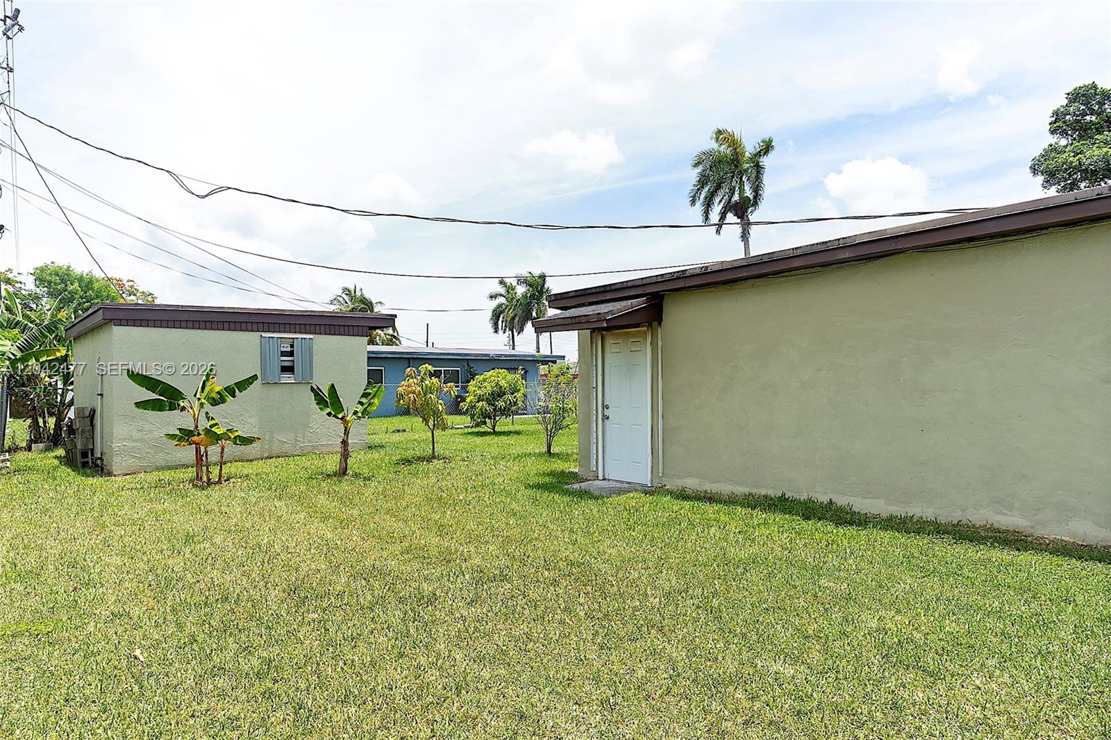 A grassy backyard features two small, light-colored buildings with a white door and palm trees — HOMESTEAD, FL