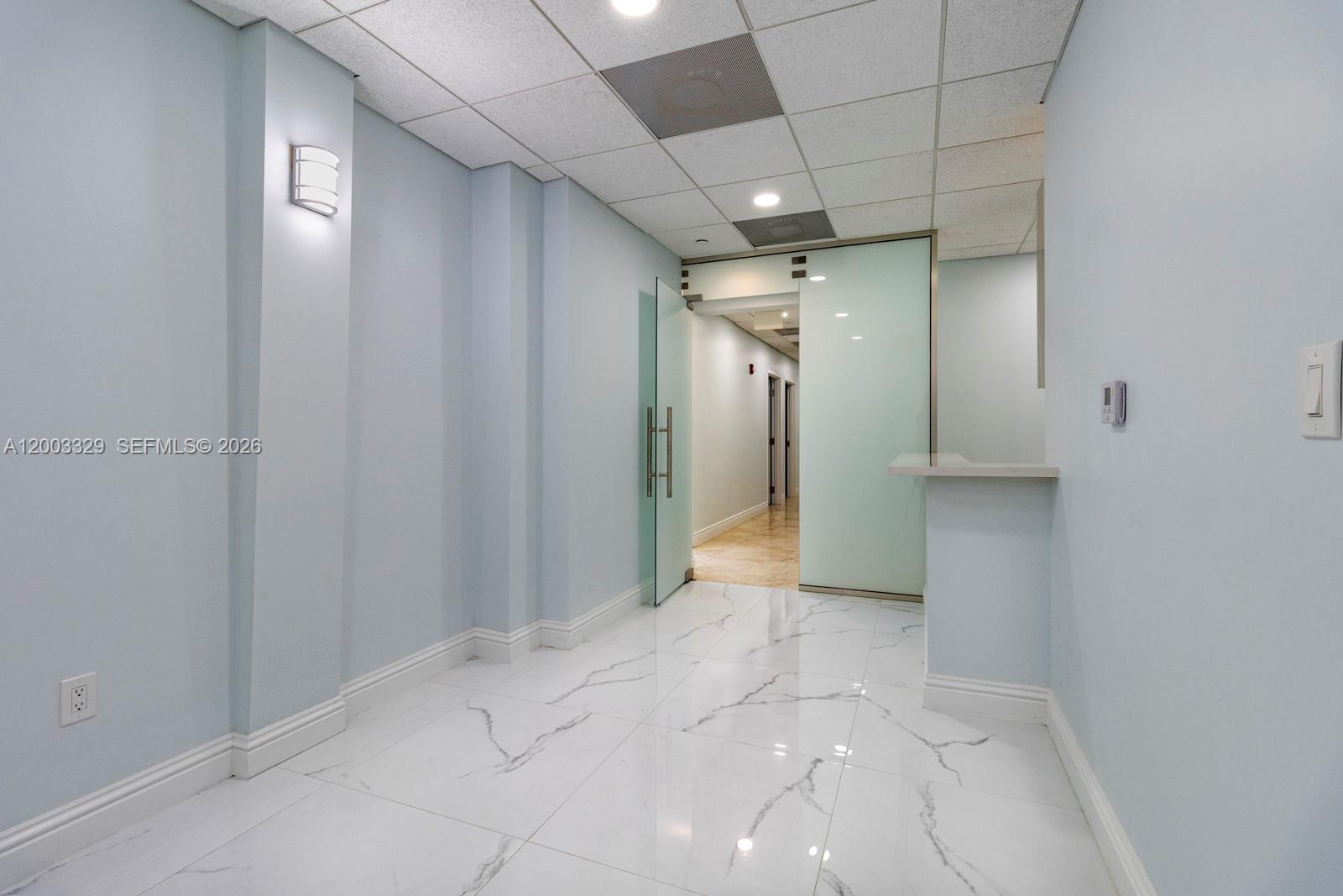 A hallway with light blue walls, white marble-patterned tile flooring, and a frosted glass door — 6141 Sunset Dr # 102, South Miami FL 33143, SOUTH MIAMI, FL