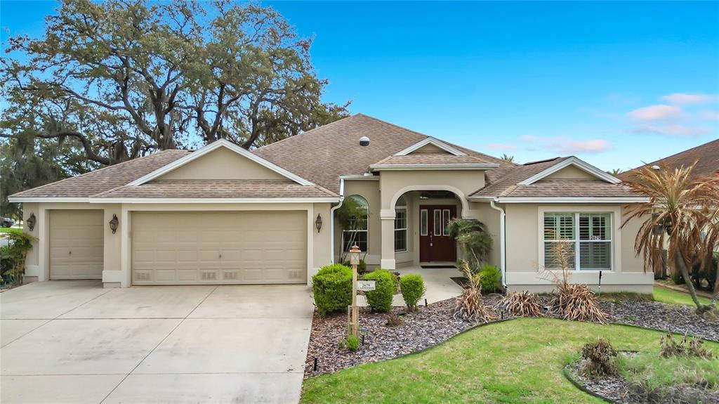 A single-story suburban house features a tan stucco exterior, a three-car garage, and a landscaped yard — 2679 LANTANA AVENUE, THE VILLAGES, FL