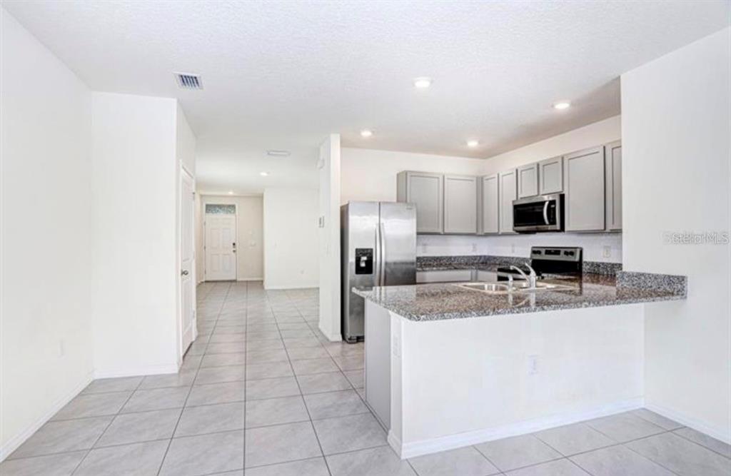 A kitchen with gray cabinets, granite countertops, and stainless steel appliances opens to a hallway — 31210 PENNY SURF LOOP, WESLEY CHAPEL, FL