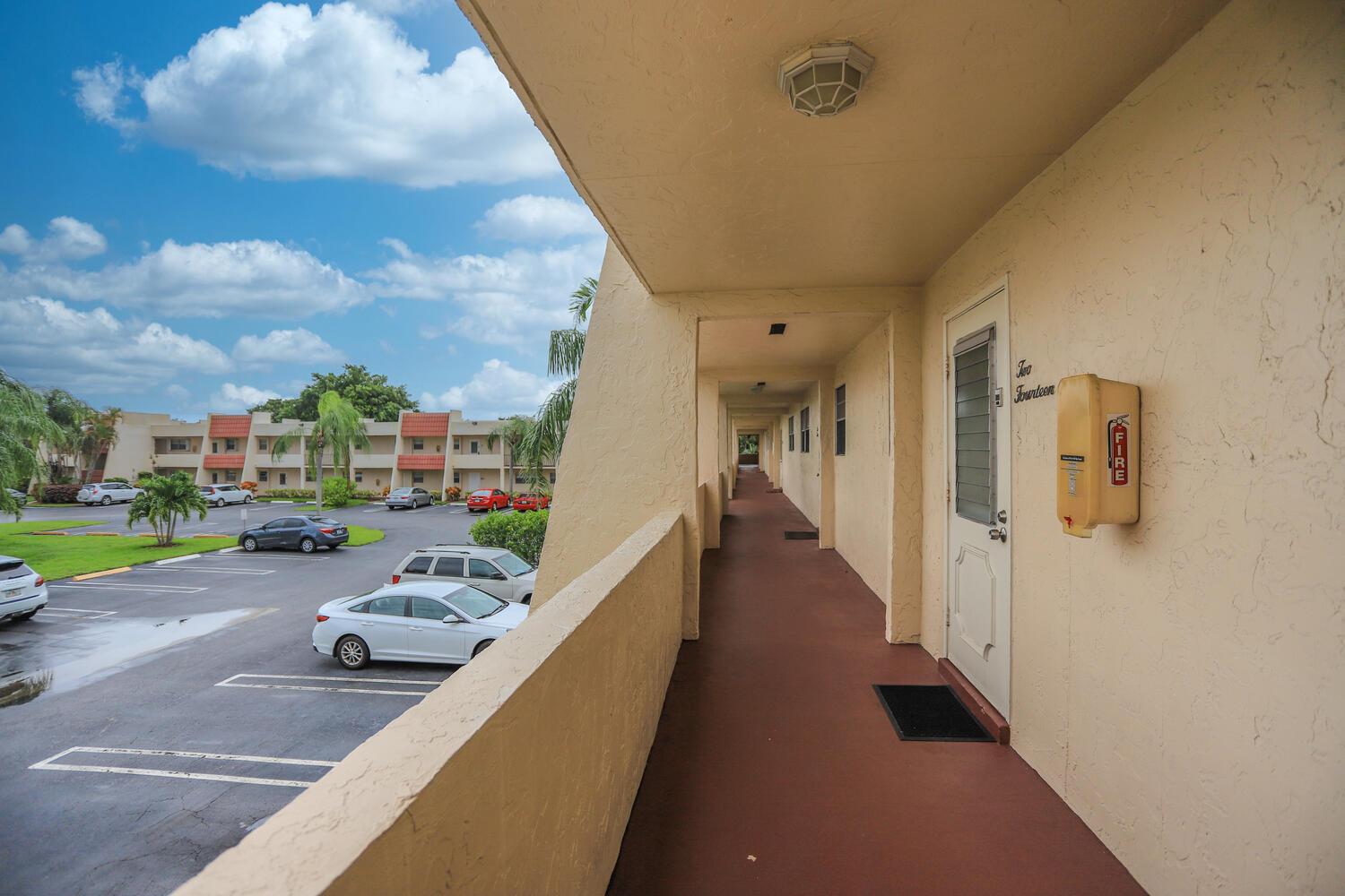An outdoor apartment walkway overlooks a parking lot with several parked cars under a blue sky — 2500 Fiore Way # a, Delray Beach FL 33445, DELRAY BEACH, FL