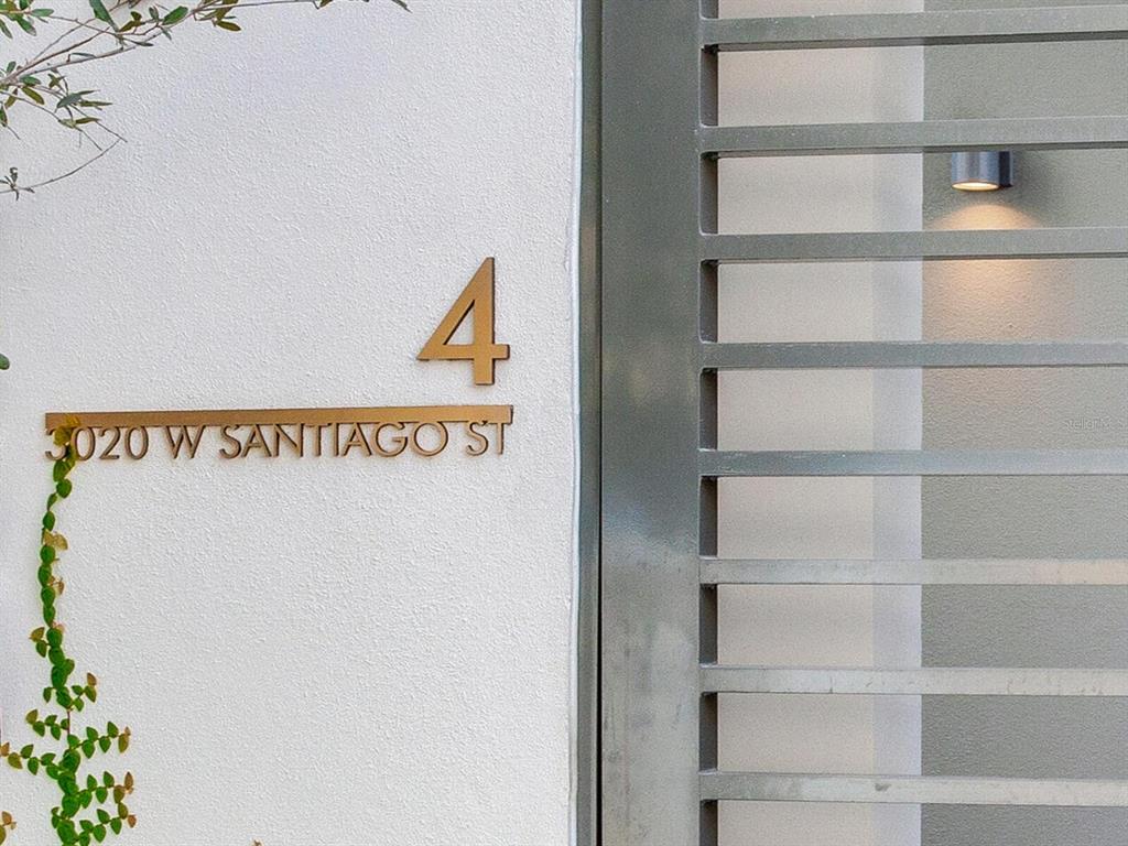 A modern metal house number sign is mounted on a white exterior wall beside a gate — 3020 W SANTIAGO STREET UNIT 4, TAMPA, FL