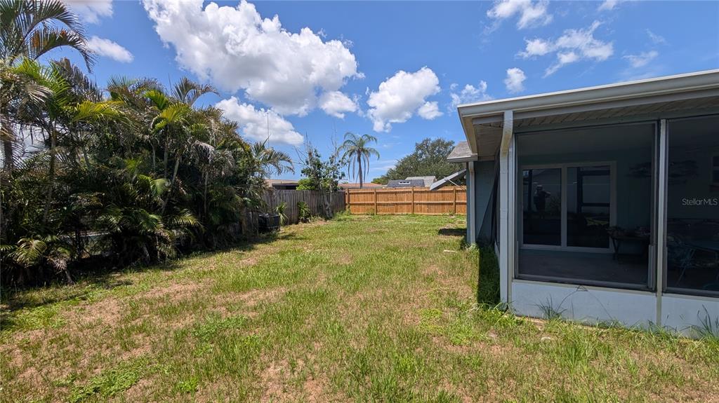 A grassy backyard features a screened-in patio and a wooden fence under a blue sky — 3452 CHAUNCY ROAD, HOLIDAY, FL