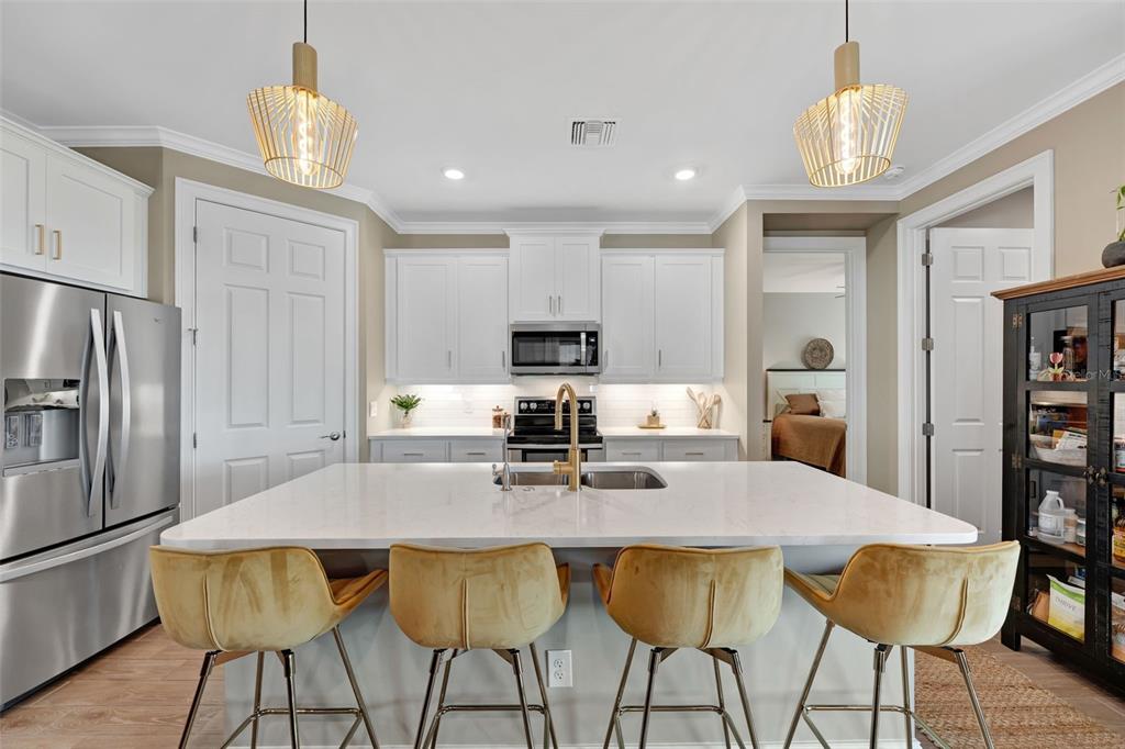 A kitchen features a white quartz island with four gold stools, stainless appliances, and white cabinetry — 16711 ELKHORN CORAL DRIVE, NORTH FORT MYERS, FL