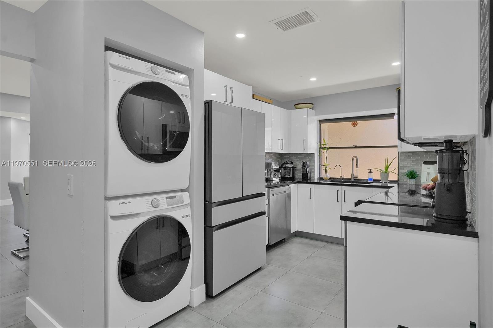 A modern kitchen features white cabinetry, black countertops, stainless steel appliances, and a stacked laundry unit — 17032 NW 63rd Pl, Hialeah FL 33015, HIALEAH, FL