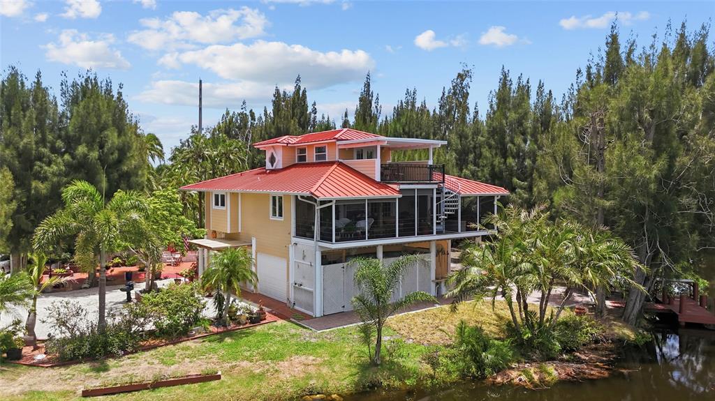 An elevated yellow house with a red metal roof sits beside a canal and trees — 10007 TAMIAMI TRAIL S, NORTH PORT, FL
