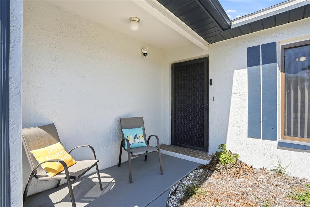 A covered front porch features two chairs, a security camera, and a screen door — 3276 MORVEN DRIVE, SPRING HILL, FL