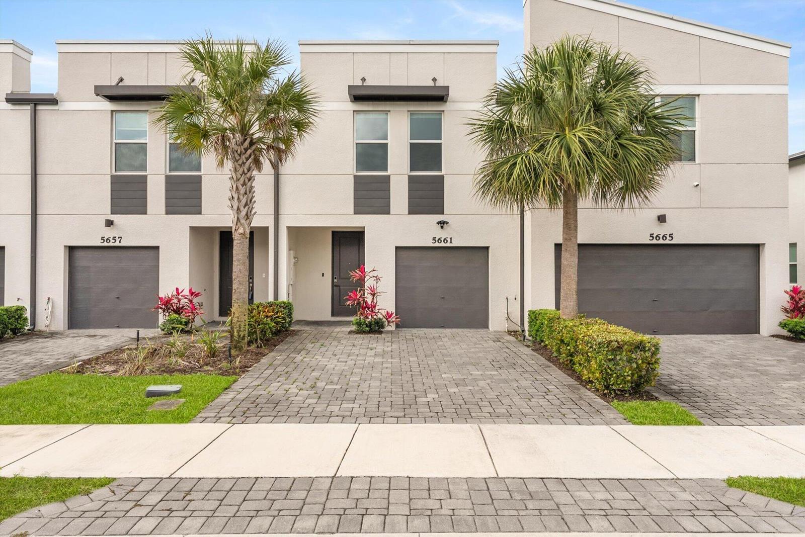 Three modern townhomes with gray garage doors and paved driveways stand under a blue sky — 5661 Mentmore Drive, Mangonia Park FL 33407, MANGONIA PARK, FL
