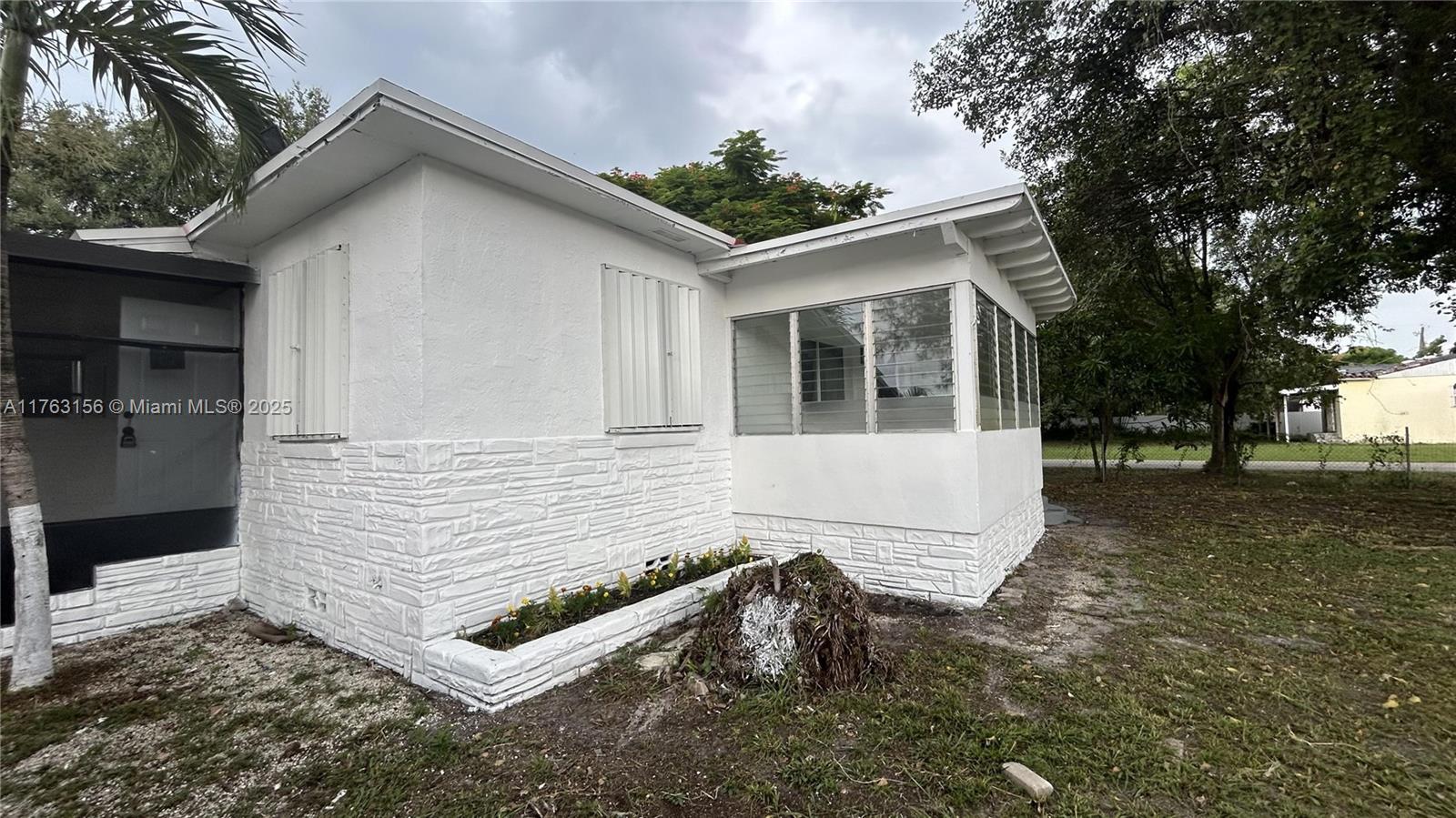 A white house exterior features a stone-textured base, louvered shutters, and a screened-in porch area — 205 Seaman Ave, Opa-Locka FL 33054, OPA-LOCKA, FL
