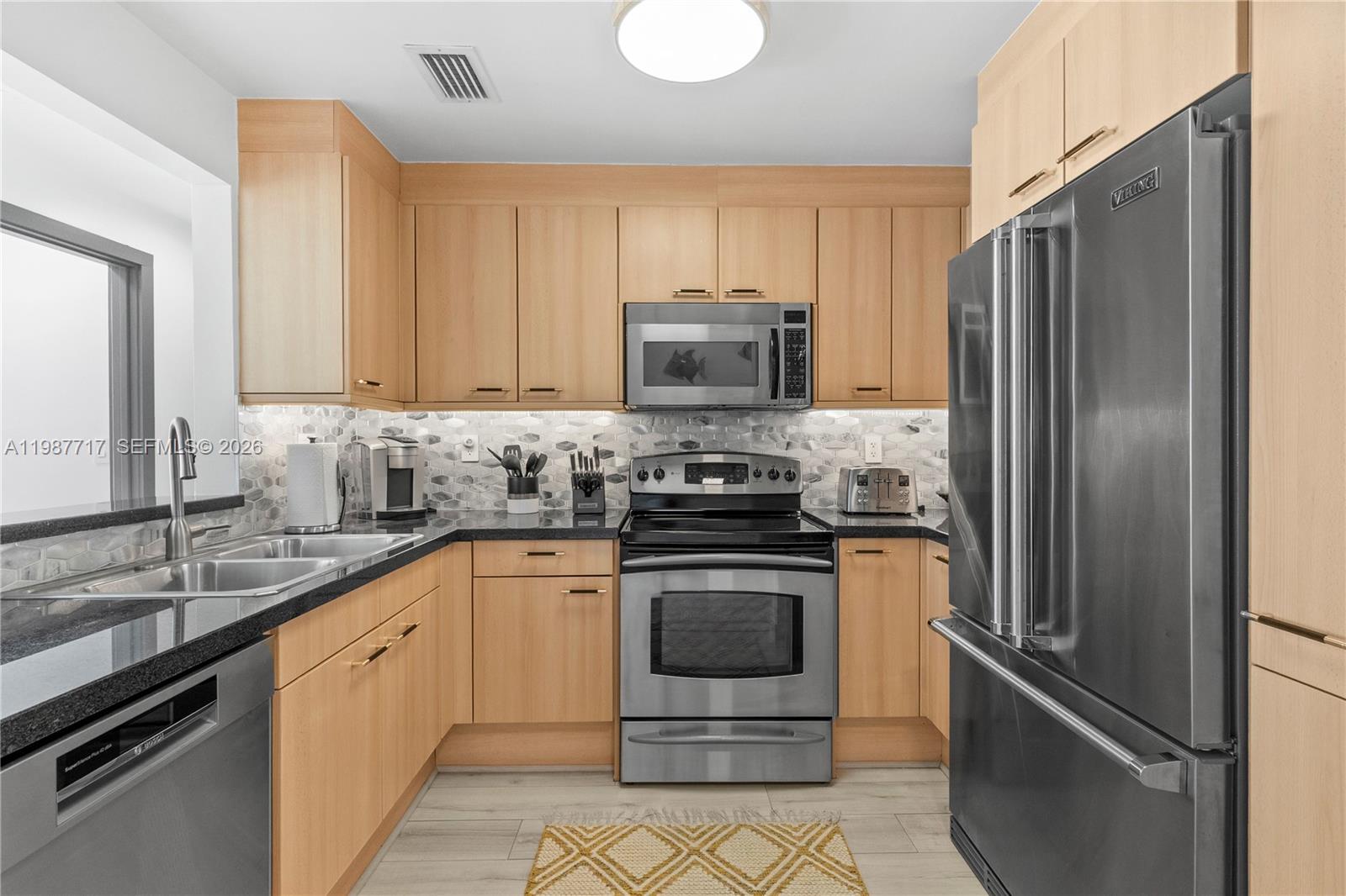 A kitchen features light wood cabinets, stainless steel appliances, a black countertop, and tiled backsplash — 225 Collins Ave # 5J, Miami Beach FL 33139, MIAMI BEACH, FL